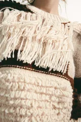 Jersey de punto con cuello redondo y manga larga, caracterizado por un diseño de parches en tonos tierra y crema. Presenta una textura variada con detalles de flecos y tejido en relieve.