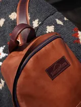 Mochila de cuero negro, con forro interior de lino y herrajes de bronce. Presenta un bolsillo frontal con cierre y un parche de cuero marrón con logo grabado.