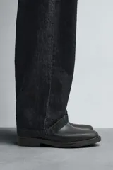 Bota corta estilo Chelsea de cuero liso negro, con elásticos laterales y tirador trasero para calce. Presenta horma redonda y suela con ligero tacón.
