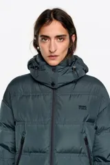 Campera tipo plumífero de largo medio con silueta en línea A, confeccionada en tejido técnico brillante. Presenta cierre frontal con cremallera, cuello subido con capucha desmontable, mangas largas con puños interiores de pelo sintético y bolsillos laterales con cremallera.