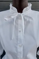 Camisa blanca de popelín con cuello redondo con volante y lazo, manga larga y bajo peplum. Cierre frontal con botones.