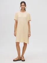 Vestido corto color amarillo claro, tipo remerón, de corte suelto con cuello redondo y mangas cortas. Presenta detalles de líneas en tono bordó en el cuello y en los puños.