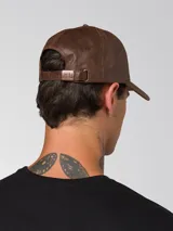 Gorro tipo gorra de béisbol con visera curva, confeccionado en cuero sintético con acabado texturizado. Presenta un pequeño aplique metálico con el logo de la marca en el lateral frontal y cierre metálico ajustable en la parte posterior.
