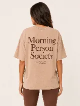 Remera oversize de algodón color tostado, con mangas cortas y cuello redondo. Presenta un gran estampado en la espalda con el texto en marrón oscuro "Morning Person Society" y una leyenda adicional.