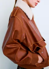 Chaqueta estilo aviador de cuero sintético marrón, con cuello de corderito sintético blanco, bolsillos frontales con solapa, cierre de cremallera y puños y cintura de canalé elástico.