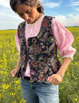 Chaleco estampado estilo tapicería con diseño floral multicolor sobre base negra, escote redondo y cierre frontal con botones metálicos.