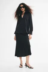 Blusa negra de tela brillante, con cuello clásico, escote en V con botones frontales, mangas largas con puños sencillos y bajo redondeado.