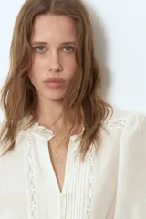Vestido túnica corto color blanco marfil, con escote en V y cuello de volados. Presenta mangas cortas abullonadas, botonadura frontal parcial y detalles de alforzas e inserciones de encaje en el frente. Cuenta con costuras fruncidas en la cadera y ribete de volado en el bajo.