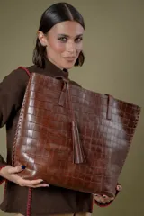 Bolso tipo tote de cuero vacuno con textura de reptil, color marrón. Presenta detalles de pasamanería en los bordes laterales y una borla de flecos de cuero colgante. Cuenta con cierre superior.