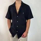 Camisa de manga corta con cuello cubano, confeccionada en tejido texturizado de color negro. Presenta cierre frontal con botones y un pequeño logo bordado en el pecho.