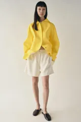 Cazadora impermeable de acabado engomado en color amarillo, con capucha ajustable, cierre frontal con botones a presión, bolsillos laterales y mangas largas ajustables.