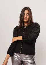 Blusa negra de tejido de punto acanalado, con cuello redondo y mangas largas abullonadas de tela semitransparente. Presenta una hilera vertical de pequeñas cuentas plateadas en el frente.