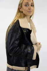 Campera oversize de cuero sintético negro con interior y detalles de corderito en tono tostado. Cuenta con cuello amplio, cierre frontal metálico cruzado, y tiras ajustables con hebilla en las mangas y la cintura.