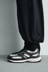 Zapatilla deportiva tipo running con diseño de paneles superpuestos en combinación de materiales. Presenta cordonera curvada y suela con volumen de diseño irregular.