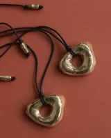 Collar con dije de forma irregular y hueco en el centro, color dorado, suspendido de un cordón negro.