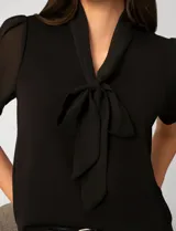 Blusa de color blanco con escote en V y detalle de lazo frontal. Presenta mangas cortas de gasa con terminación en puño.