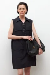 Vestido negro sin mangas con cuello camisero y cierre frontal con cordones. Presenta dos bolsillos con solapa a la altura del pecho y dos bolsillos laterales a la altura de la cadera.