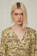 Vestido corto color amarillo con estampado abstracto en marrón, escote en V cruzado, mangas largas abullonadas con puños elásticos y falda con volantes.