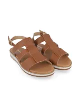 Sandalia de cuero color camel con diseño de tiras cruzadas y detalle de flecos en la parte superior. Cuenta con cierre de hebilla metálica ajustable en el tobillo y suela plana de goma con plataforma bicolor.