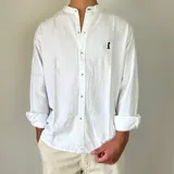Camisa de lino blanca con cuello mao y cierre frontal de botones. Presenta un pequeño bordado oscuro en el pecho y mangas largas con puños doblados.