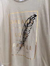 Remera de algodón color beige con corte relajado y diseño asimétrico, más larga en la parte posterior. Presenta un estampado frontal rectangular con letras doradas, una pluma ilustrada y detalles de pedrería decorativa.
