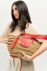 Bolso tipo canasto de paja con detalles en cuero rojo, incluyendo un bolsillo externo con solapa y cierre con botón a presión, y correa regulable.