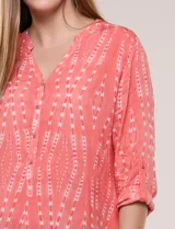 Blusa turquesa de silueta suelta confeccionada en tejido liviano, con escote en V, pequeña cartera frontal con botones decorativos y mangas 3/4 con presilla para arremangar. Presenta patrón estampado geométrico tipo tie-dye y ruedo curvo.