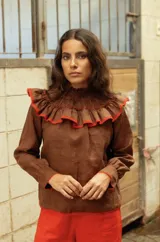 Camisa de lino color marrón, con cuello alto fruncido y detalle de volados en el pecho con borde en color rojo. Presenta mangas largas con puños abotonados.