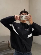 Campera deportiva negra con cuello alto, cierre frontal completo y franjas blancas en las mangas. Presenta estampados en color rosado en el pecho, incluyendo la inscripción "Only F.A.N.S." y gráficos de palmeras con estrellas.