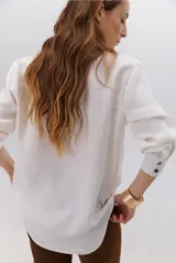 Camisa beige de manga larga con botones marrones y cuello clásico. Presenta un detalle de costura dorada a lo largo de la botonera.