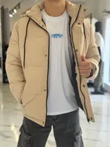 Campera puffer impermeable color beige con capucha desmontable con cordones negros, cierre frontal con cremallera y solapa con botones a presión.