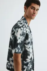 Camisa de manga corta con cuello abierto, tapeta frontal con botones y bajo recto. Presenta un estampado floral con efecto desgastado en tonos blanco y negro.