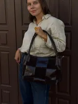Cartera de hombro confeccionada en cuero y gamuza con diseño de damero, combinando paneles de texturas lisas y aterciopeladas. Presenta una manija trenzada y detalles de costuras artesanales en los laterales.