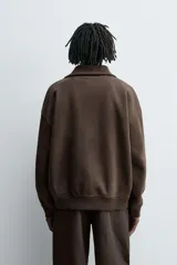 Sudadera de corte relajado color marrón oscuro, con cuello subido y cierre frontal de cremallera. Presenta manga larga, bolsillo frontal tipo canguro y acabados en rib en puños y ruedo.