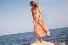 Vestido largo con estampado caleidoscópico en tonos rosa, rojo y verde. Presenta tirantes finos, escote en V, corte imperio y falda con volantes. Incluye detalle de trenzas en el pecho y la cintura que se anudan en la espalda descubierta.