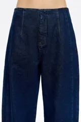 Pantalón de denim azul oscuro, corte barrel (tobillero con volumen en la cadera y estrechamiento hacia el tobillo). Presenta tiro medio, cierre frontal con corchete oculto y cremallera, y tiras ajustables con hebilla en la espalda.