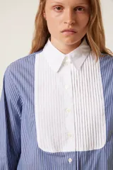Camisa de popelina a rayas azules y blancas, de corte recto, con cuello y puños blancos. Presenta un babero plisado blanco.