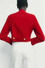 Cazadora de estilo militar en color rojo, con cuello redondo, manga larga y cierre frontal mediante alamares con botones dorados. Presenta trabillas con botón en los hombros y detalle de costuras en los puños.