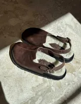 Zueco de cuero color chocolate con interior forrado en piel sintética blanca. Presenta una tira trasera ajustable con hebilla metálica y suela de goma negra.