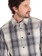 Camisa de manga larga con estampado a cuadros en tonos beige, gris y negro. Presenta cuello clásico, cierre frontal con botones y un bolsillo en el pecho con el logo de la marca.