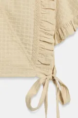 Top crop de escote en pico y manga corta con volados. Presenta un diseño cruzado que se ajusta mediante una lazada lateral y cuenta con una textura de cuadrícula sutil en toda la prenda.