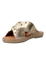 Sandalias tipo ojota con tiras cruzadas en cuero metalizado dorado, decoradas con tachas metálicas y hebilla lateral. Presentan una base con detalle de yute trenzado alrededor del contorno.