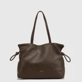 Cartera tipo tote de hombro, color marrón oscuro, con diseño de textura granulada y detalles de nudos laterales. Cuenta con doble asa larga y logo de la marca grabado en la parte inferior frontal.