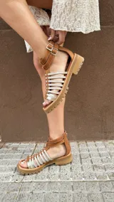 Sandalias bajas de cuero color beige con tiras horizontales plateadas y cierre con hebilla al tobillo.