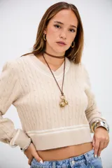 Sweater de punto trenzado en color beige, con cuello en V y detalles de rayas blancas en el cuello, puños y ruedo. De corte ajustado y manga larga.
