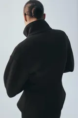 Chaqueta negra de manga larga con cuello subido, bolsillos delanteros y cierre frontal con botones ocultos bajo solapa.