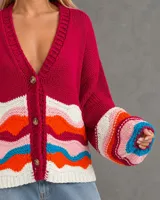 Cardigan tejido de punto en color fucsia, con escote en V y cierre frontal de botones. Presenta un diseño de ondas multicolor en la parte inferior y en los puños, con mangas largas de corte holgado.
