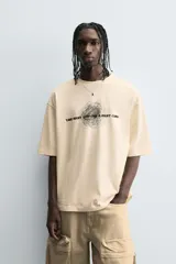 Camiseta de algodón de corte relajado, color beige claro, con cuello redondo y manga corta. Presenta un estampado central de garabatos negros con texto en mayúsculas que dice "TAKE RIGHT STEP FIND A RIGHT CORD".