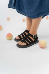 Sandalia estilo gladiadora de color beige, confeccionada en material gamuzado sintético. Presenta múltiples tiras anchas que sujetan el pie, ajuste lateral con hebilla metálica y suela plana antideslizante de color marrón.