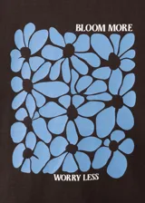 Camiseta marrón de algodón con cuello redondo y manga corta, con estampa frontal azul de flores y texto "Bloom More Worry Less".